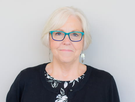 Close-up portrait of a smiling person with short gray hair, wearing blue-framed glasses, a black cardigan over a white top with a black floral pattern, and dangling earrings. The background is a light gray solid color.