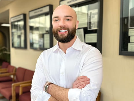 Man with a beard wearing a white shirt smiles while standing in a hallway with photo frames behind him. 