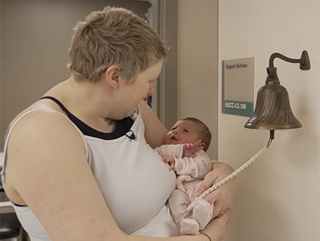 Profile of a woman with short blond hair looking lovingly at her tiny infant. They are standing near a bell mounted on the wall at The University of Kansas Cancer Center.