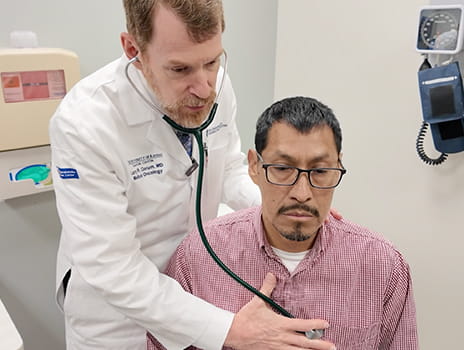 Healthcare provider using a stethoscope to listen to a seated patient’s chest during a medical exam in a clinical room.