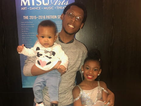 Stephon Fisher, a young man with glasses, is pictured smiling while holding a child in one arm with his other arm around the shoulder of his daughter.