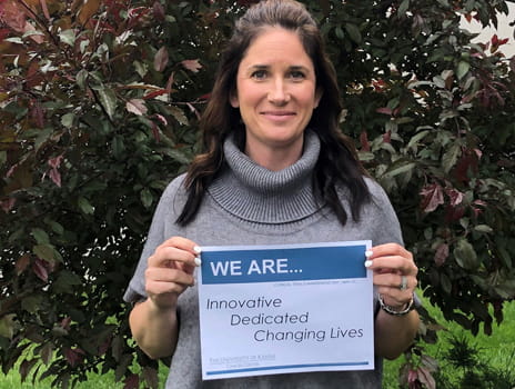woman with long brown hair holds a sign that says "we are innovative, dedicated, changing lives"