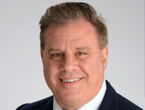 Portrait of a smiling person wearing a dark suit and tie against a light gray background.