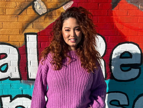 Woman with long, curly brown hair stands in front of mural smiling