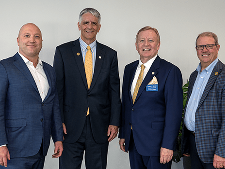 Four people dressed in business suits smile and pose in front of a blank wall.